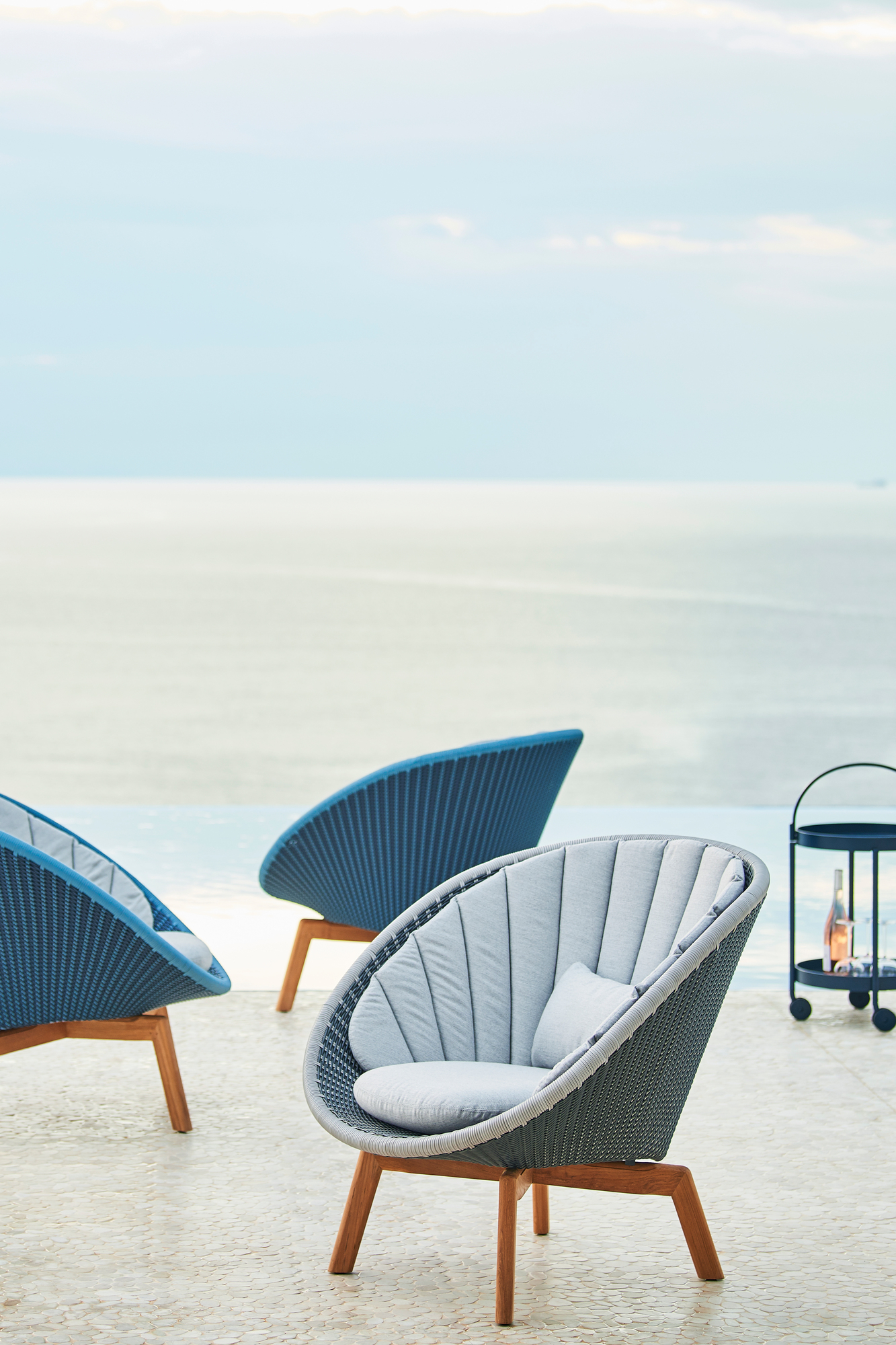 Cane-Line Peacock Loungesessel aus Rattan mit grauen Polstern und Holzbeinen im Außenbereich.