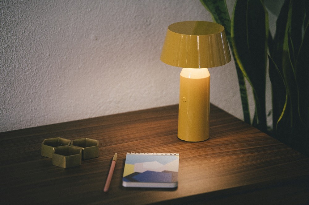 Gelbe Bicoca Akkuleuchte von Marset auf einem Holztisch mit Notizbuch und Stift.