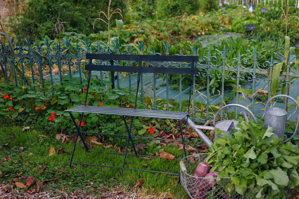 Fermob Bistro Klappbank für zwei Personen im Garten, neben Gießkannen und Gemüsekorb.