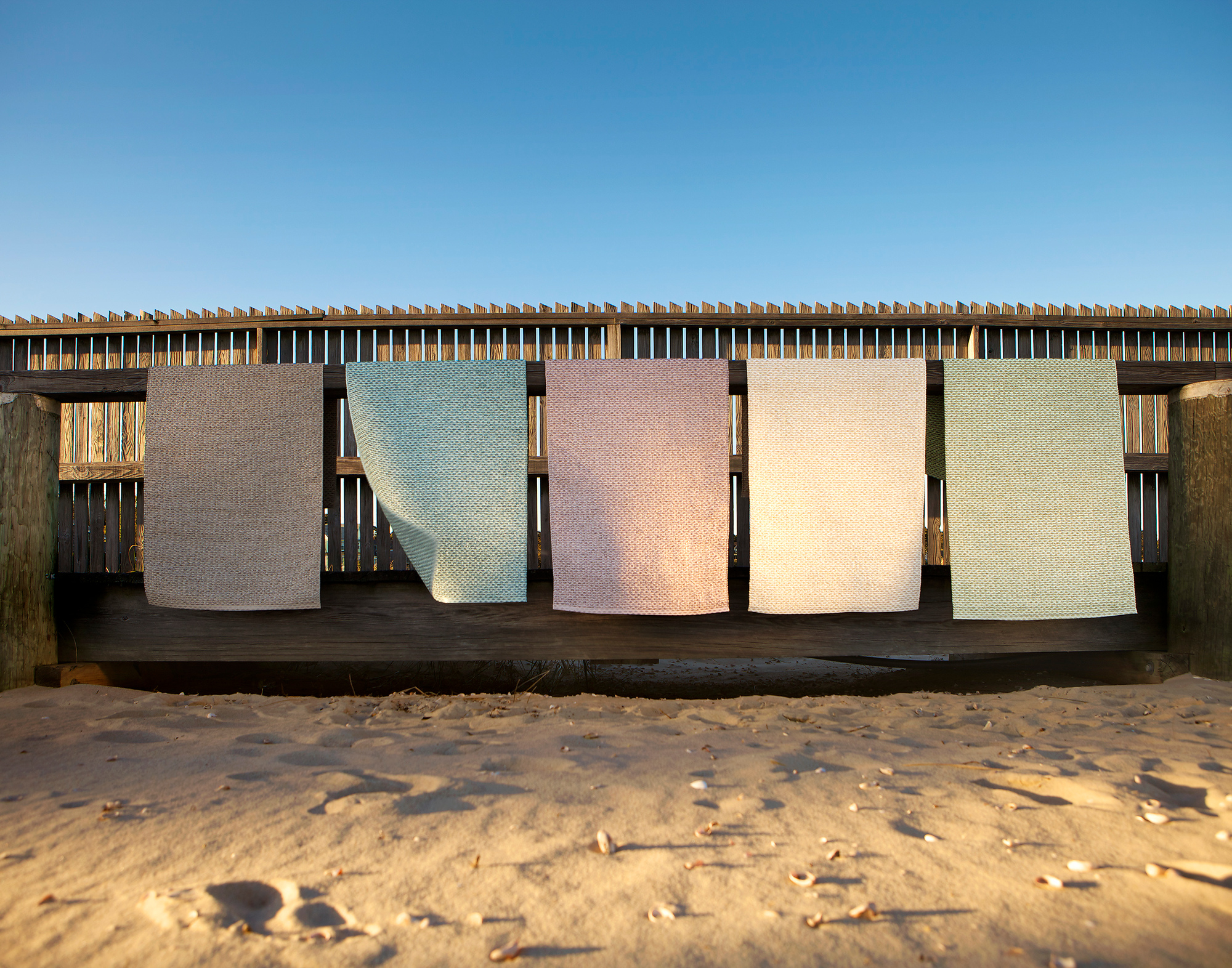Svea Kunststoffteppich von Pappelina, 70x240 cm, in verschiedenen Farben auf einem Holzgeländer am Strand.