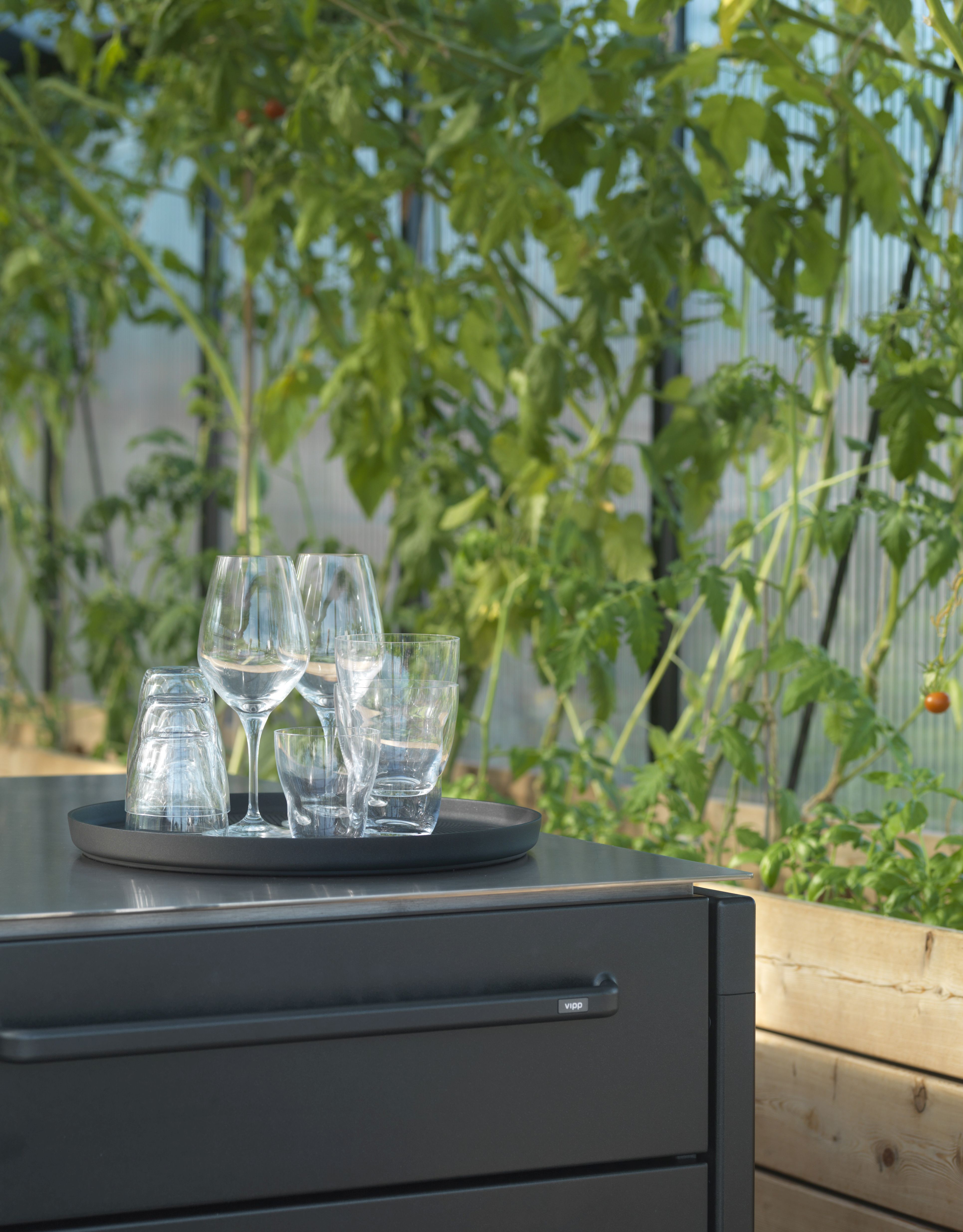 Schwarzes Vipp Tablett mit Gläsern auf einem Outdoor-Küchenschrank vor einem Gewächshaus mit Tomatenpflanzen.