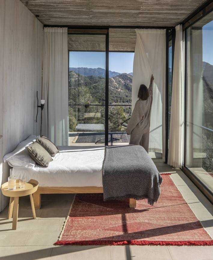 Schlafzimmer mit Bett, rotem Teppich und Frau, die weiße Vorhänge vor Panoramafenster mit Bergblick zuzieht.
