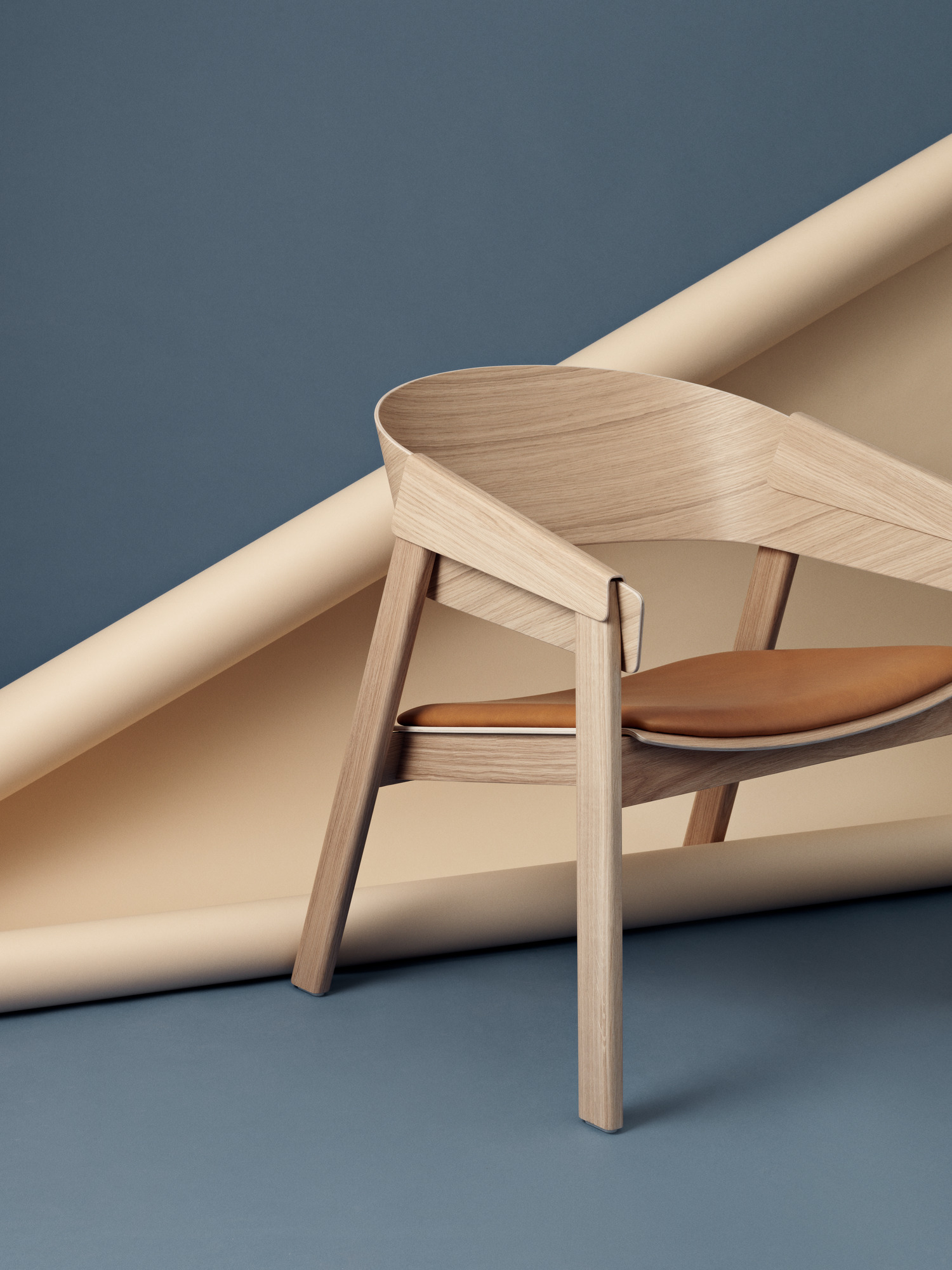 Muuto Cover Lounge Chair Stuhl aus Eichenholz mit braunem Ledersitz vor blauem Hintergrund.