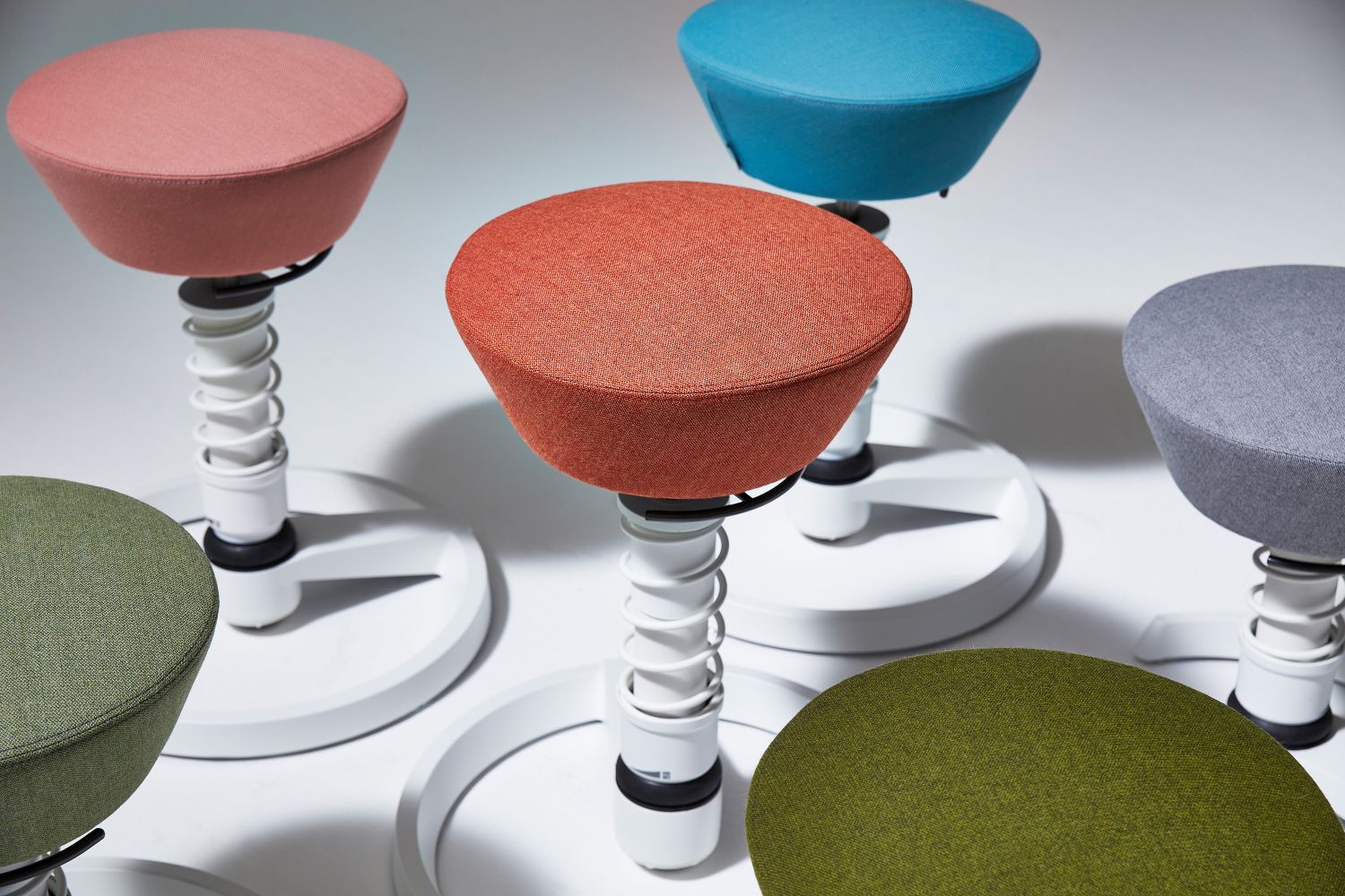 Mehrere bunte, moderne Stehhocker mit Textilbezug und weißem Federfuß für ergonomisches Sitzen.