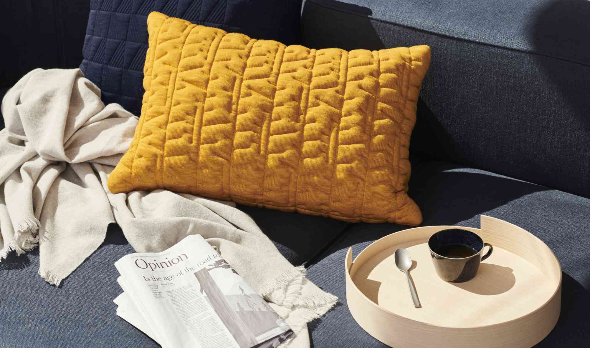 Detailaufnahme des Fritz Hansen Alphabet Sofas mit gelbem Kissen, Decke, Zeitung und Getränk auf einem Tablett.