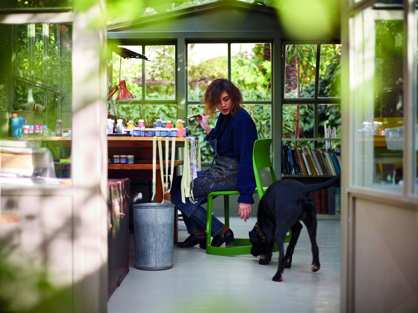 Frau sitzt auf grünem Vitra Tip Ton RE Stuhl in Atelier, malt und wird von Hund beobachtet.