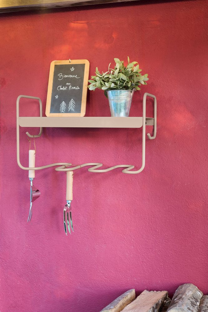 Beiges Fermob Picolino Wandregal für drinnen und draußen an pinker Wand mit Deko und Gartengeräten.