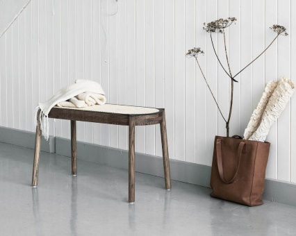 Holzbank mit Rattan-Sitzfläche und weißer Decke, neben brauner Ledertasche und Trockenblumen vor weißer Wand.