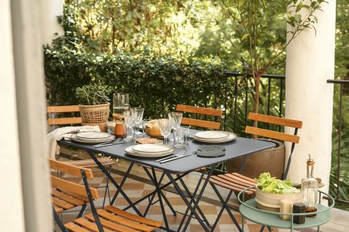 Fermob Bistro+ Gartentisch mit Stühlen auf einer Terrasse, gedeckt mit Geschirr und Speisen für ein Essen im Freien.