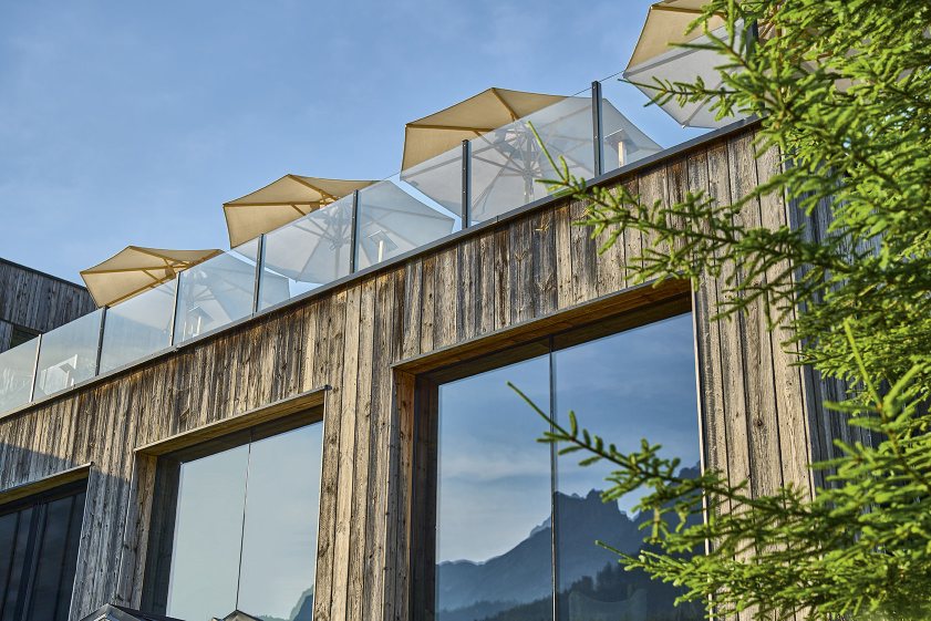 Weishäupl Klassiker Sonnenschirme auf dem Dach eines Holzgebäudes mit Glasgeländer, reflektiert in großen Fenstern.
