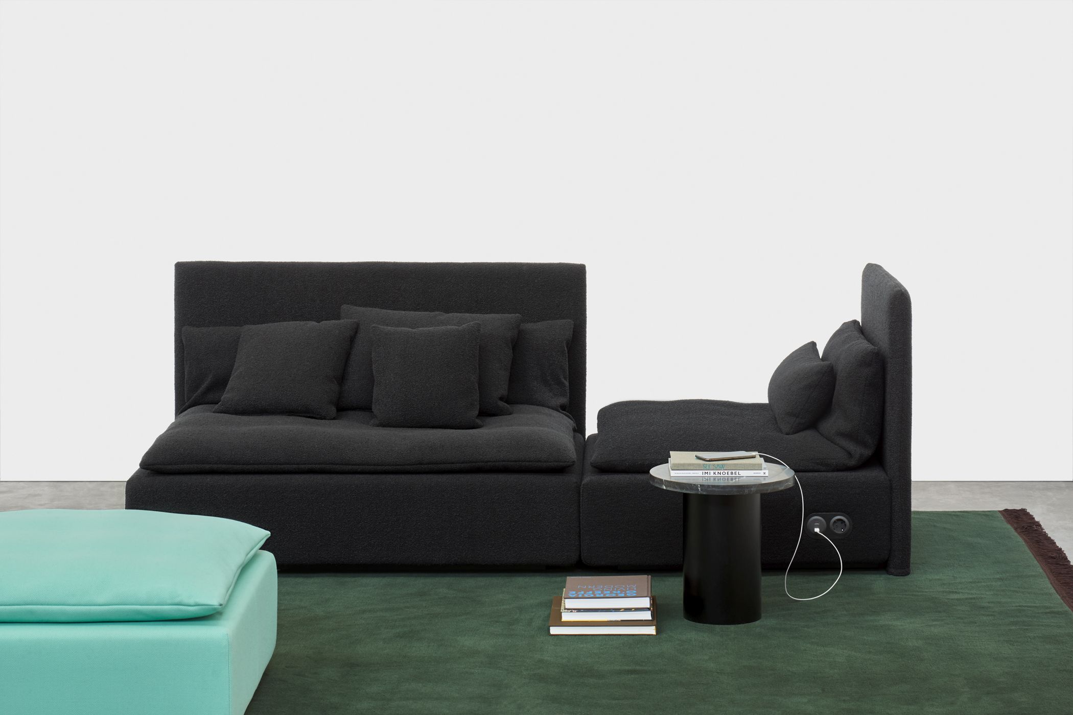 Enoki CT09 Beistelltisch von e15 neben modularem Sofa und türkisfarbenem Hocker auf grünem Teppich.