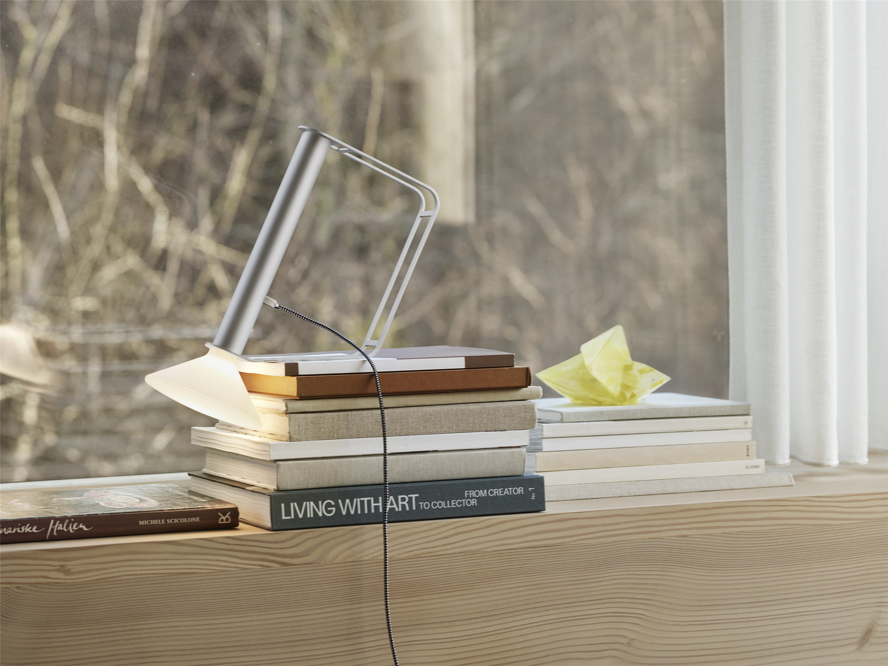 Silberne LED-Tischleuchte auf Bücherstapel vor Fenster, modernes Design für Büro und Wohnzimmer.