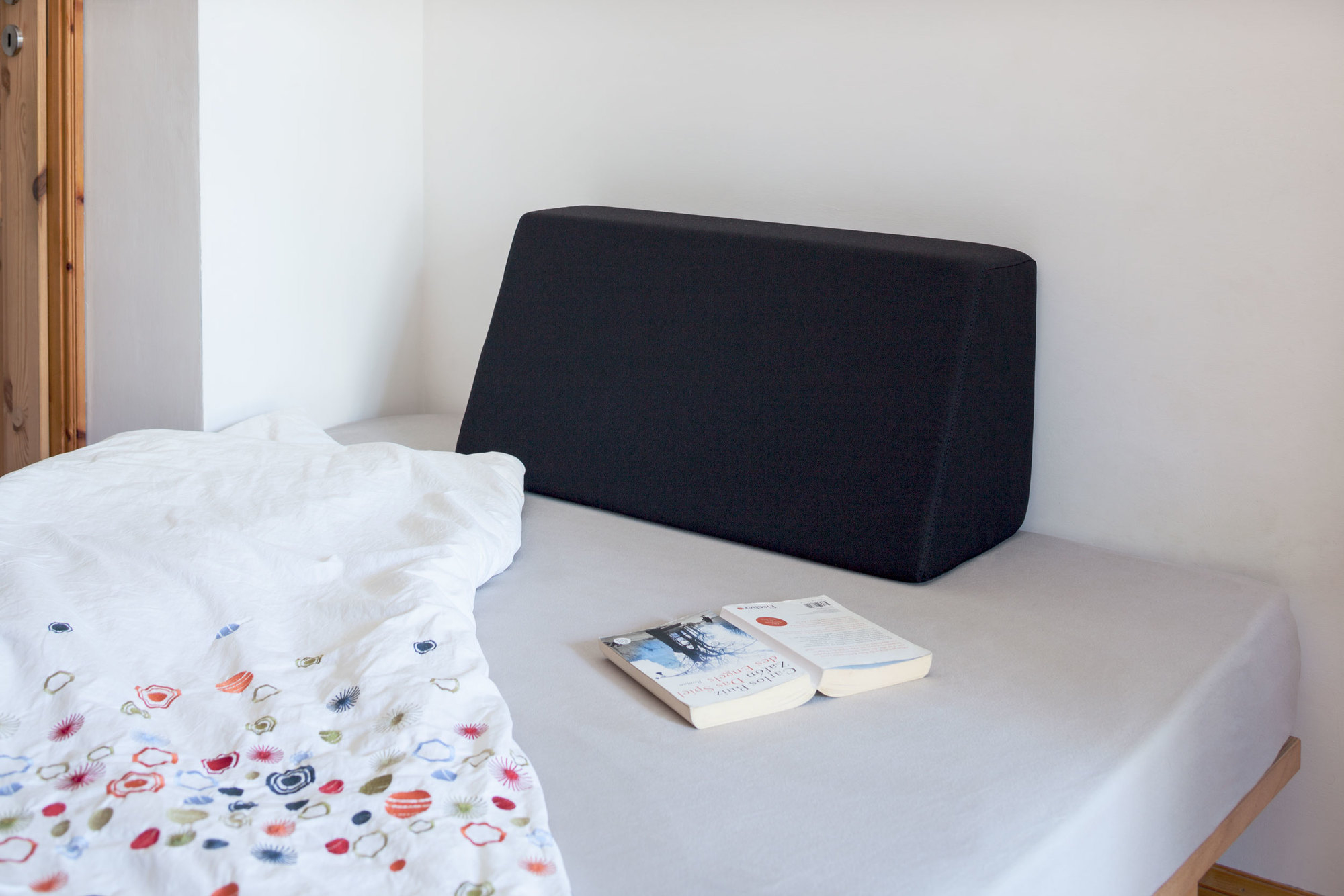 Schwarzes Tojo Lehn Rückenkissen auf einem Bett mit weißer Bettwäsche und Buch.