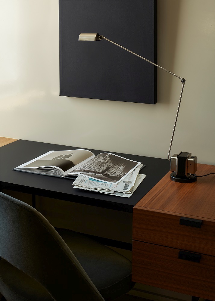 Schreibtisch mit Lampe, Magazin und Zeitungen, kombiniert mit einem Holzschrank und einem grauen Stuhl.