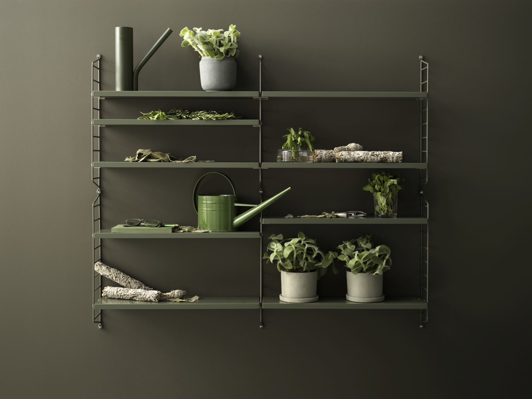 Grünes String Pocket Regal mit Pflanzen, Gießkannen und Dekoration an einer dunklen Wand.