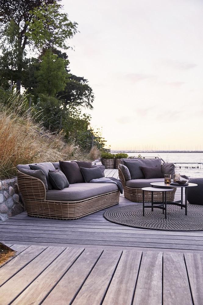 Cane-Line Basket Outdoor Sofa 2-Sitzer aus Rattan mit grauen Kissen und Beistelltisch auf Holzterrasse.