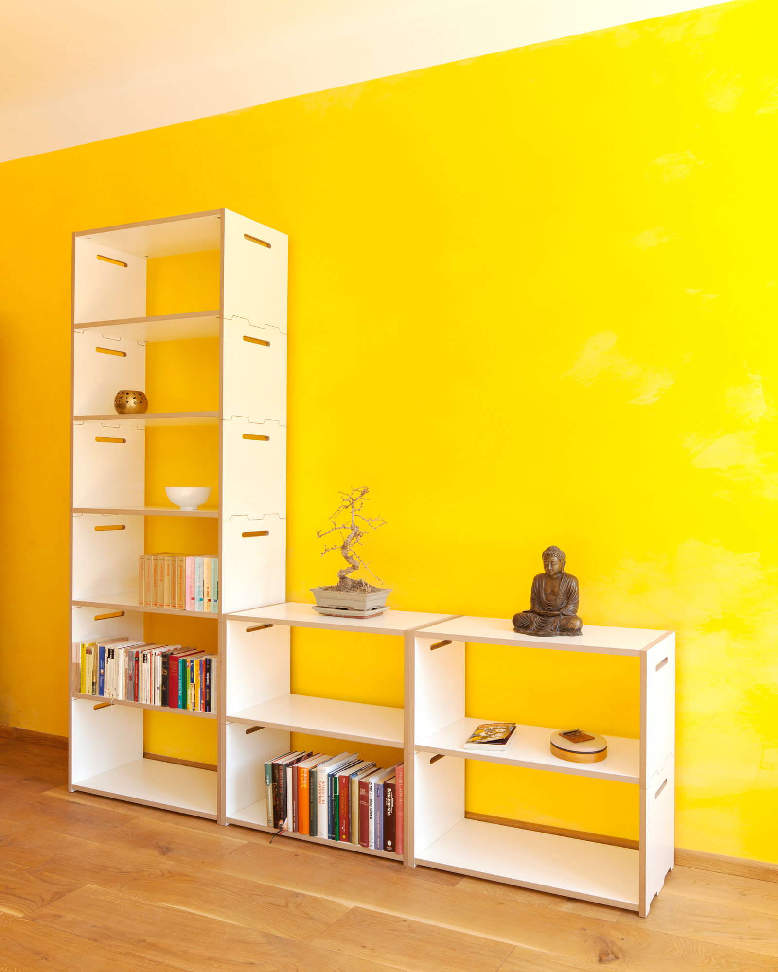 Tojo Hochstapler Regal Anbaumodul in Weiß vor gelber Wand, dekoriert mit Büchern und Deko.