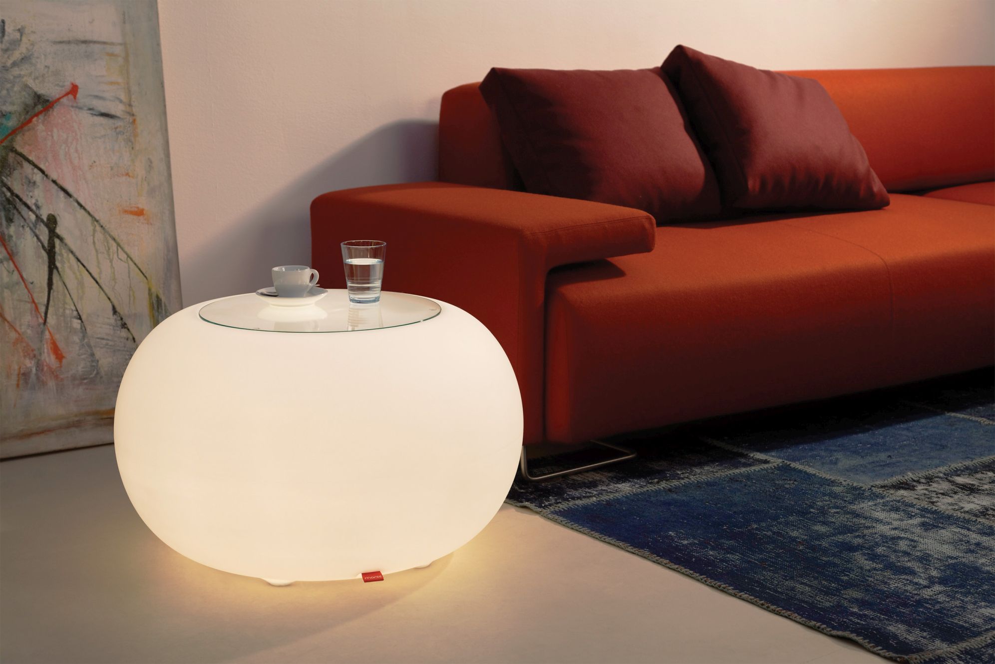 Moderne, leuchtende Kugelleuchte als Beistelltisch mit Glasplatte, Tasse und Wasserglas neben rotem Sofa.