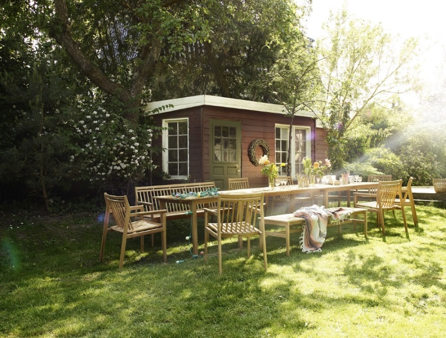 Gartenmöbel-Set aus Holz mit langem Tisch, Stühlen und Bänken vor Gartenhaus im Grünen.