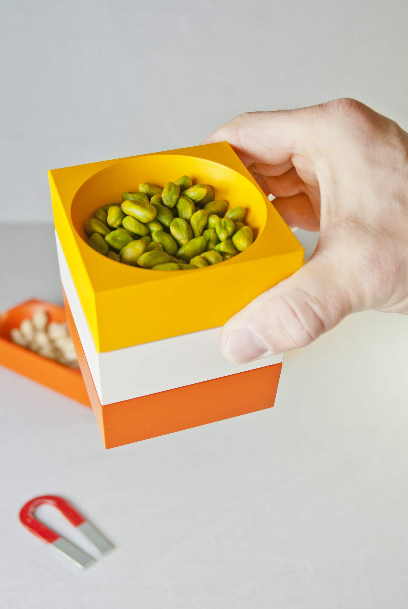 Modul 4 Magnetic Bowl Schale: Hand hält gelbe Schale mit Pistazien, gestapelt auf weißen und orangen Schalen.