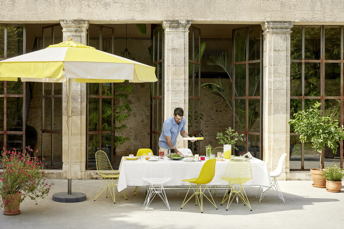 Ein Mann deckt einen Tisch mit DKR-2 Wire Chairs von Vitra in Gelb und Weiß.