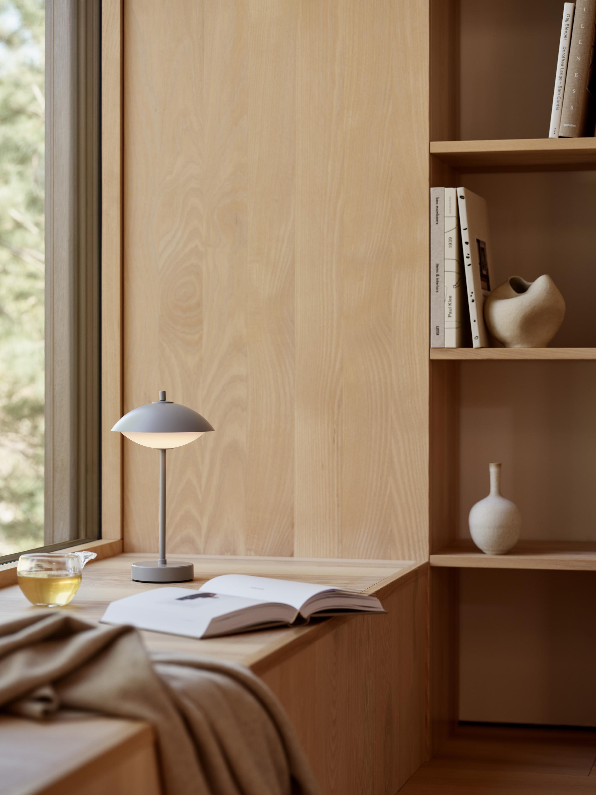 Clam Tischleuchte von Fritz Hansen in Dusk Blue auf einem Holztisch mit Buch.