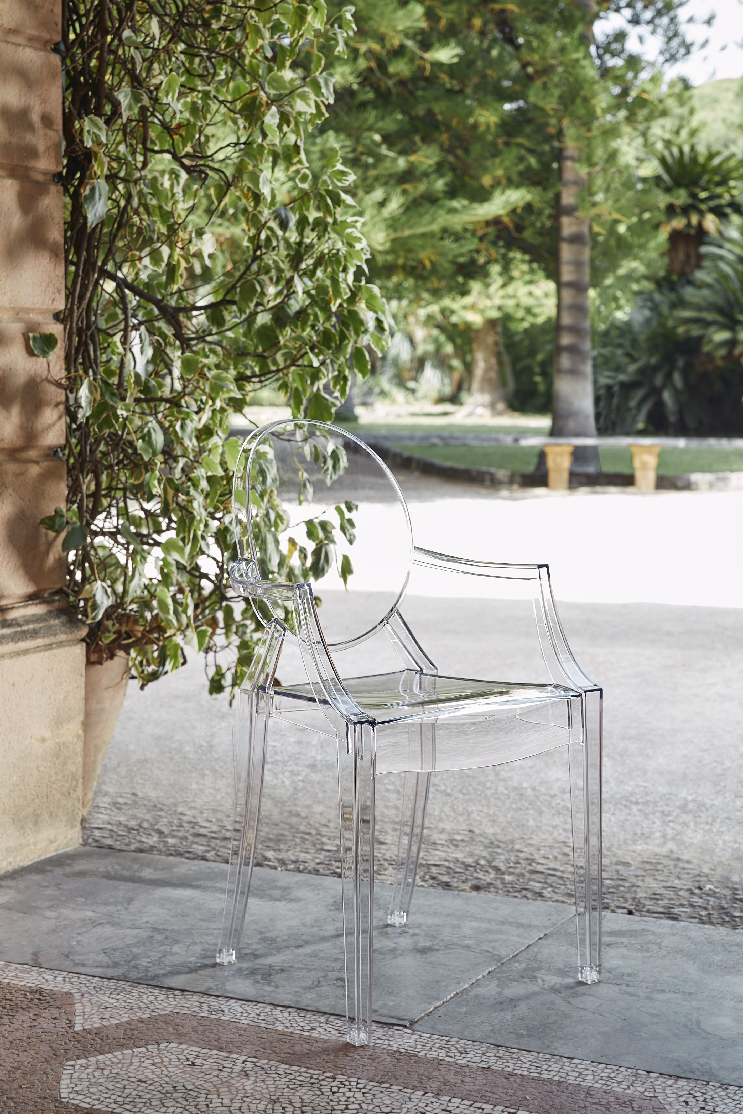 Glasklarer Louis Ghost Armlehnenstuhl von Kartell vor Grünpflanzen im Außenbereich. Designstuhl aus transparentem Kunststoff.