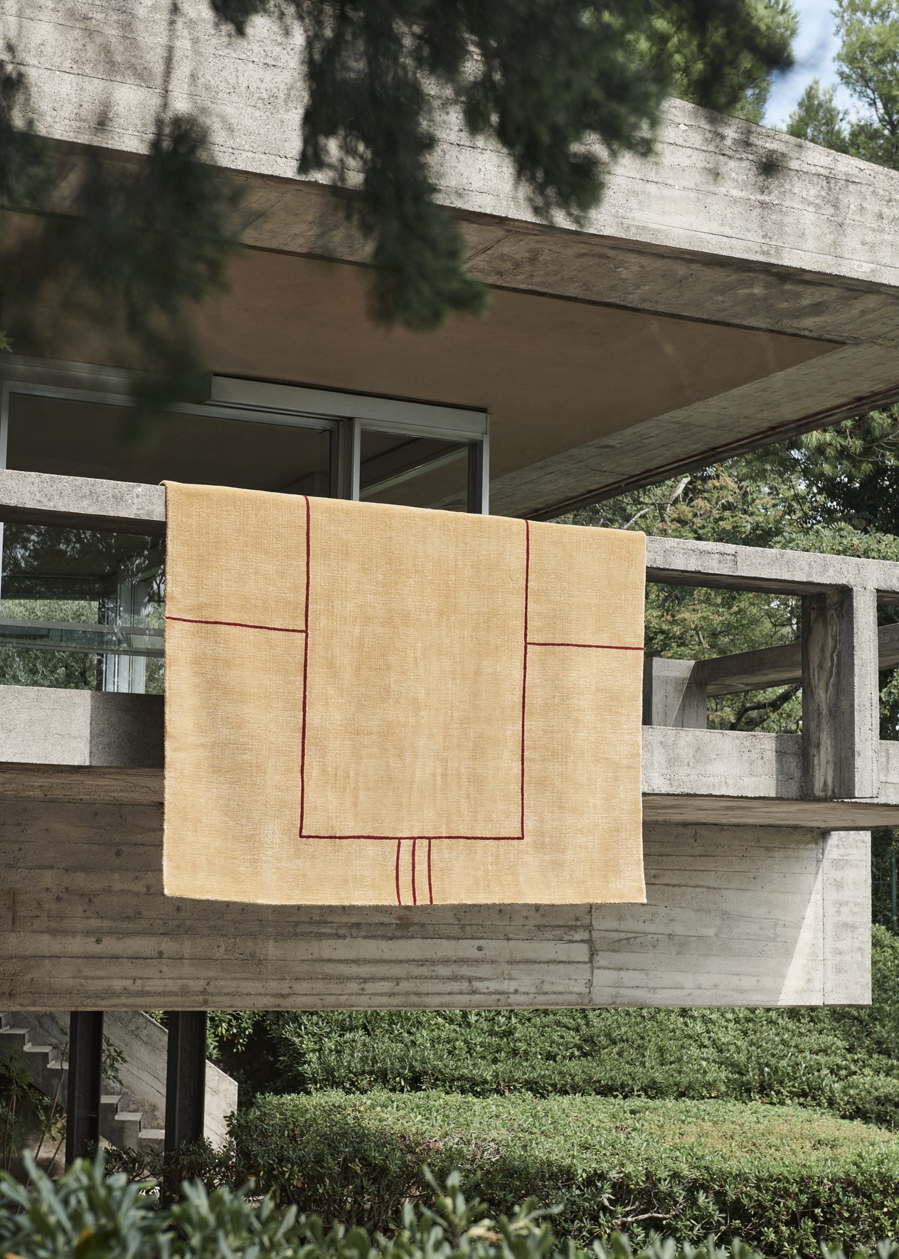 AndTradition Cruise Teppich AP11 in Beige mit rotem geometrischen Muster vor Betonfassade.