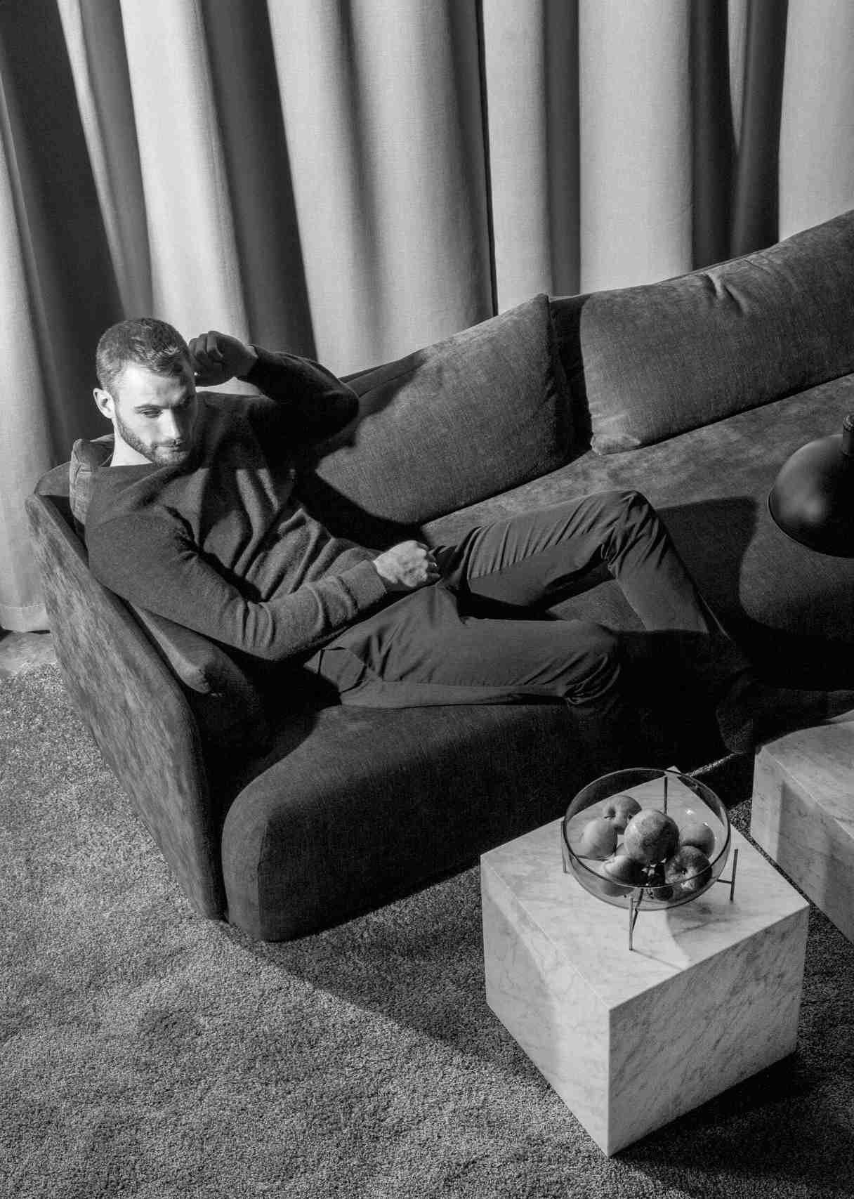 Schwarz-Weiß-Foto: Mann entspannt auf Sofa neben Audo Copenhagen Plinth Marmor Tisch mit Obstschale.