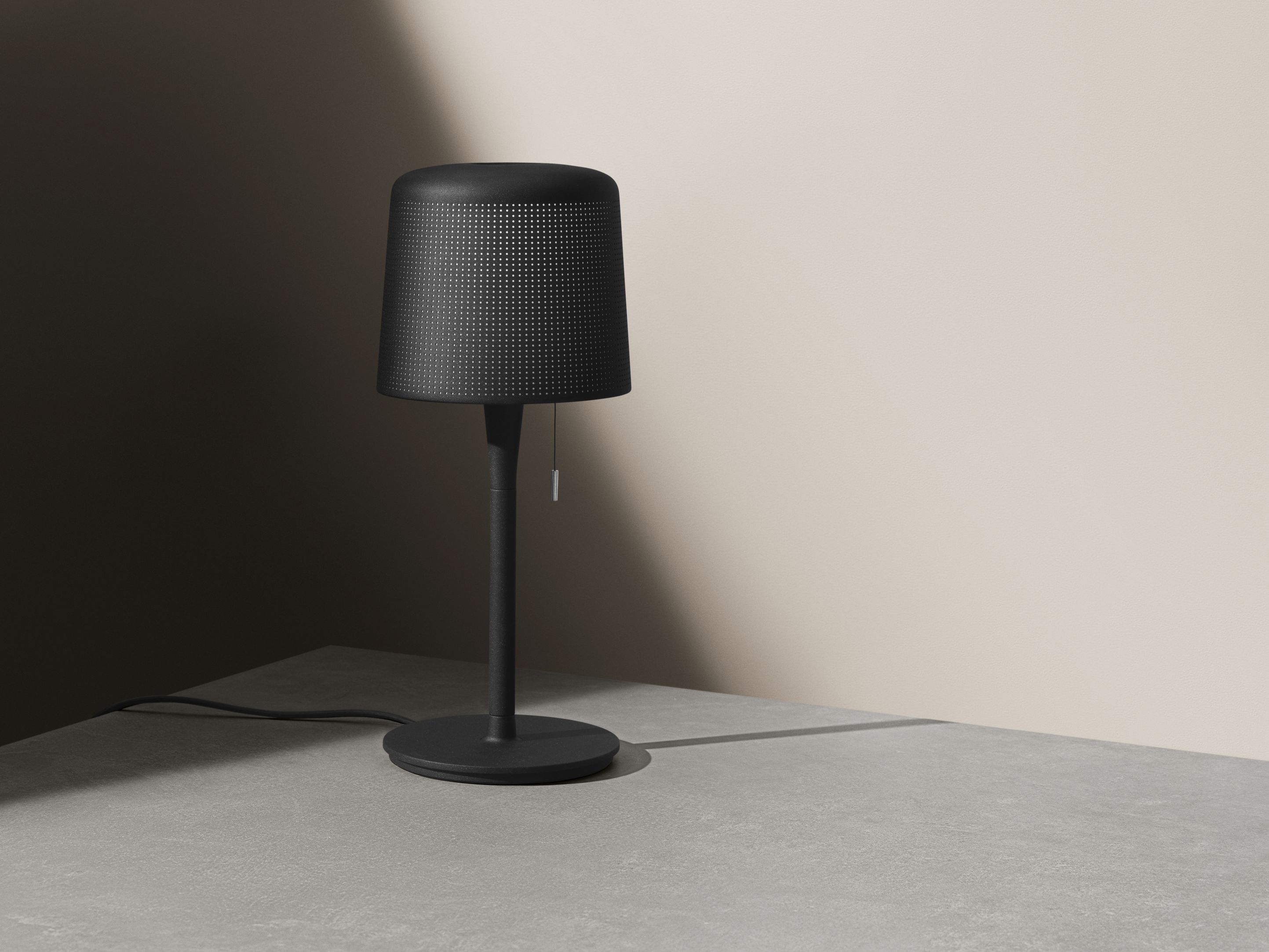 Schwarze Vipp Tischleuchte mit perforiertem Lampenschirm und Zugschalter auf einem grauen Tisch, modernes Design.