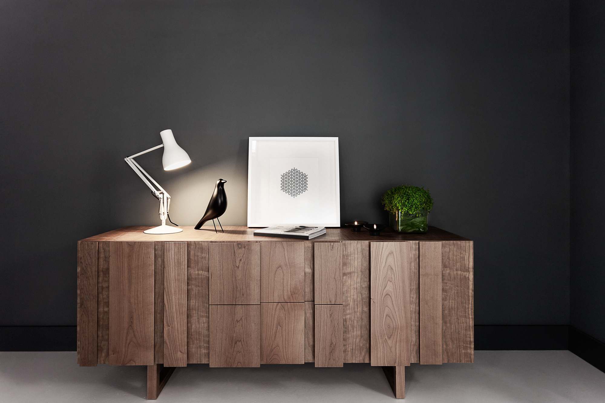 Weiße Anglepoise Type 75 Schreibtischleuchte auf Holzkommode mit Deko-Elementen vor dunkler Wand.