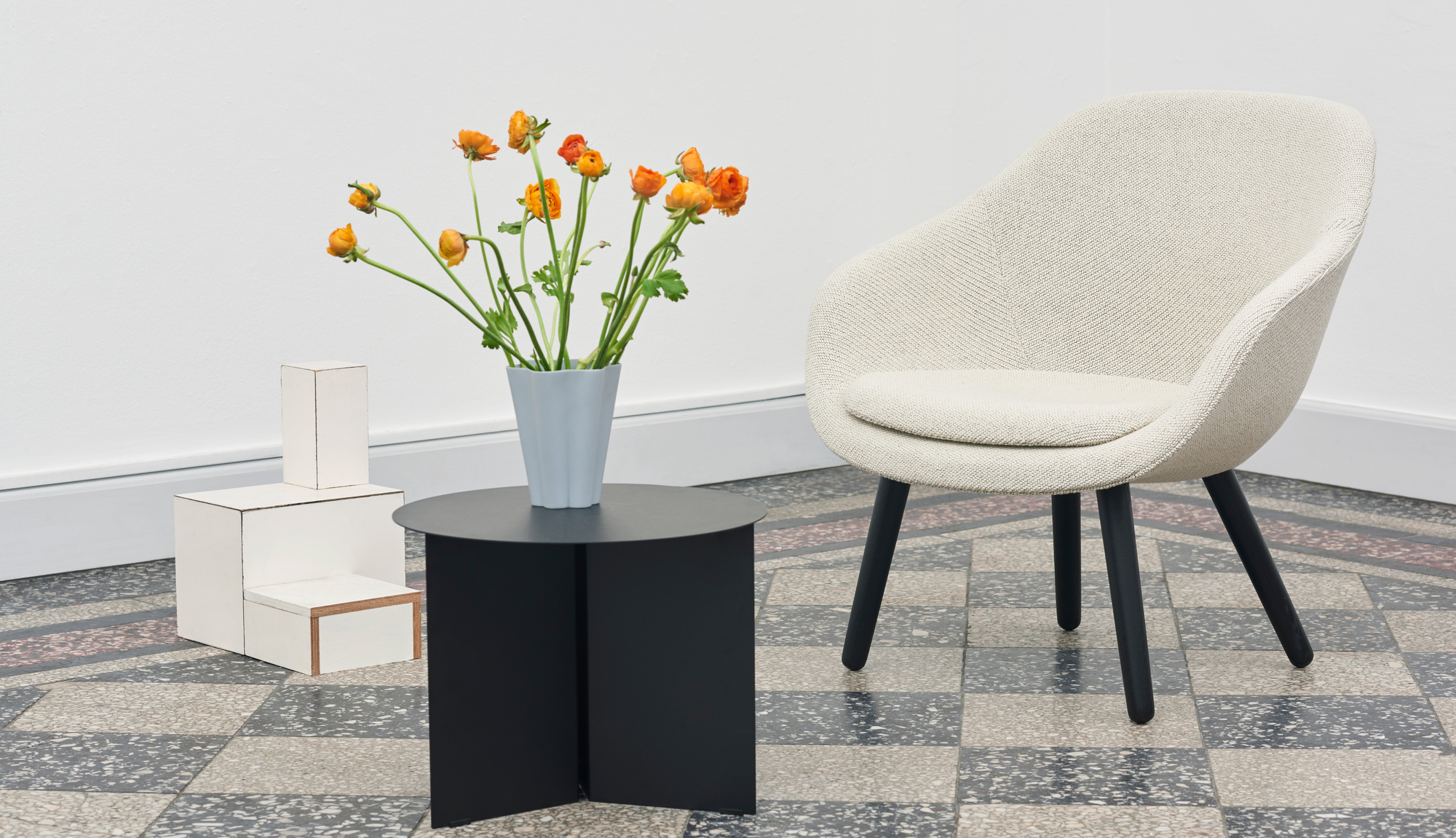 Beiger About a Lounge AAL 82 Sessel von Hay neben schwarzem Tisch mit Blumen auf Schachbrettboden.