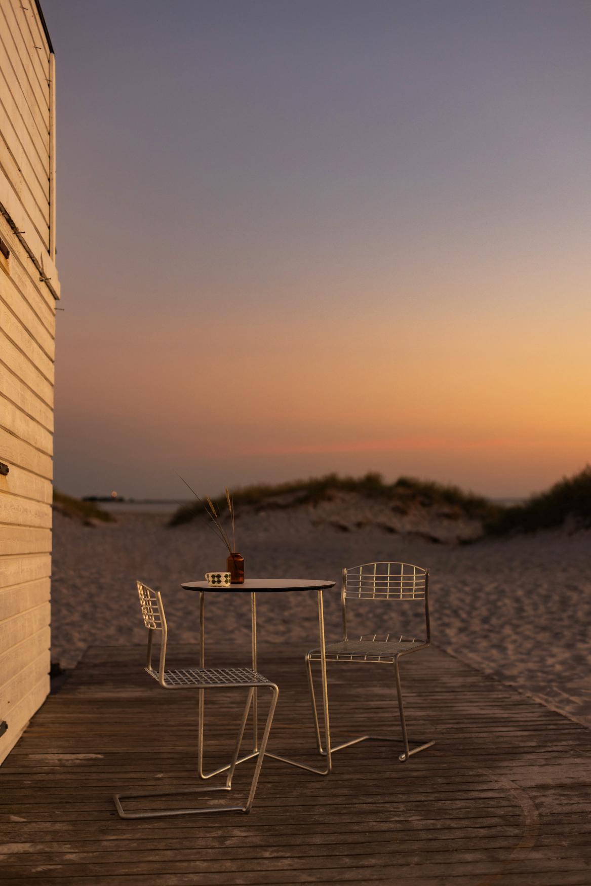 Gartenset aus Tisch und Stühlen aus Metall auf Holzterrasse am Strand bei Sonnenuntergang.