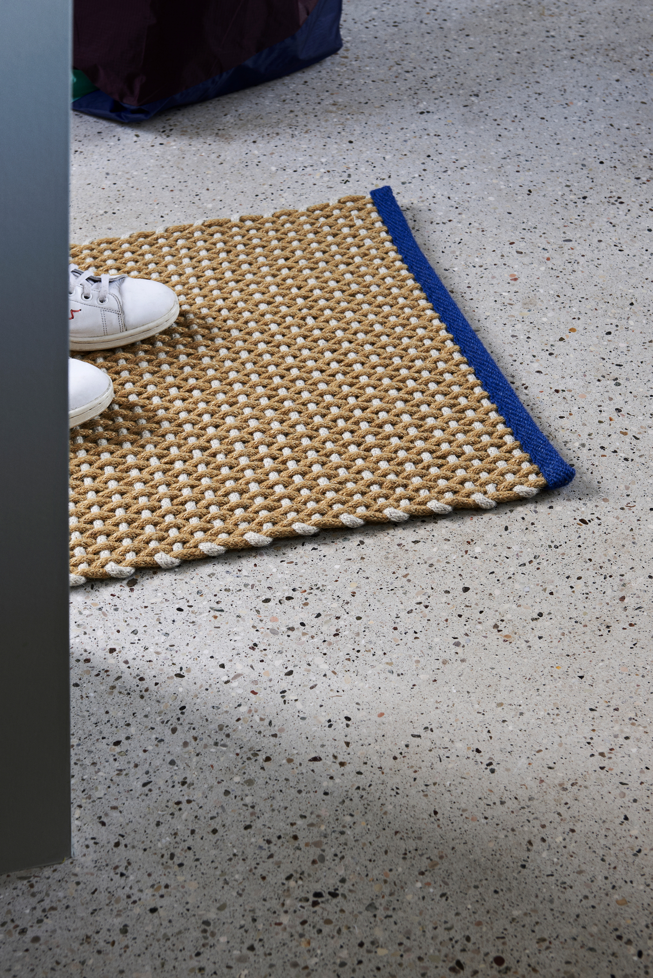 Beiger Hay Teppich mit blauem Rand und weißen Schuhen auf grauem Boden. Fußmatte für den Eingangsbereich.