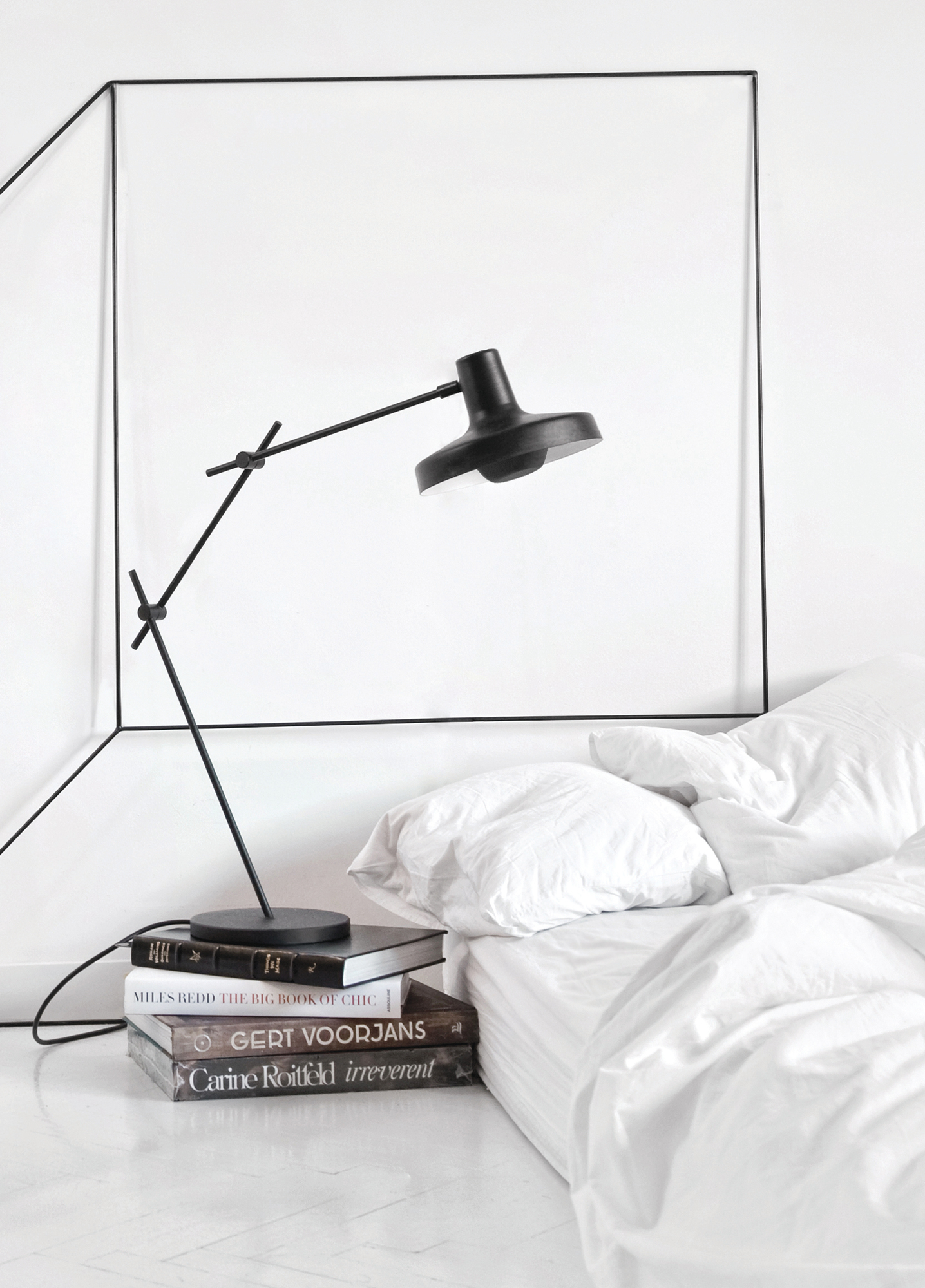 Arigato Tischleuchte von Grupa, schwarze Designlampe auf Bücherstapel neben einem weißen Bett.