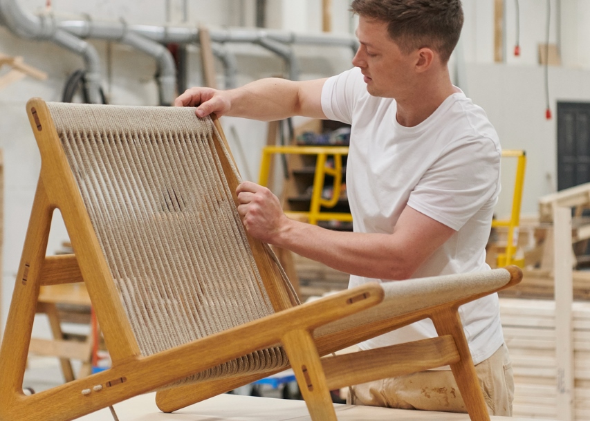 Mann montiert handgefertigten Holzstuhl mit Seilbespannung in Werkstatt. Möbeldesign, nachhaltige Produktion.