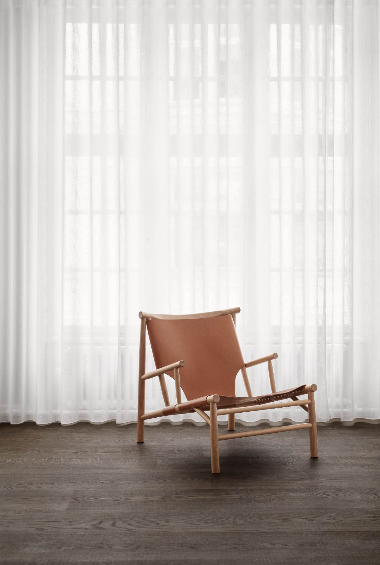 Samurai Lounge Chair Sessel von NORR11 mit hellem Holzrahmen und cognacfarbenem Leder vor weißem Vorhang.
