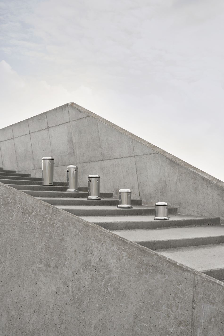 Fünf Vipp Treteimer 14L aus Edelstahl stehen auf einer grauen Betontreppe vor einer Betonwand.