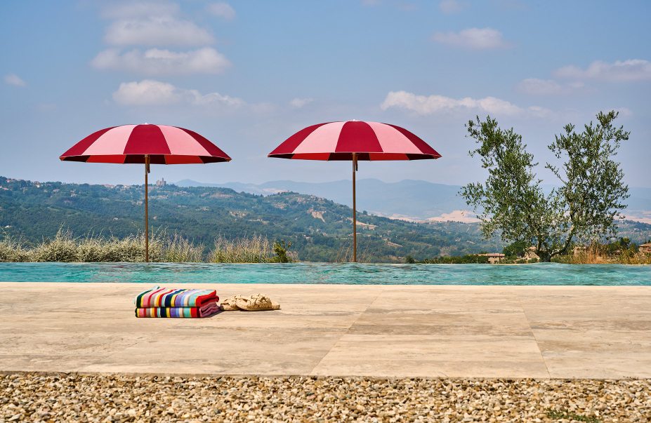 Zwei rot-weiß gestreifte Weishäupl Sonnenschirme am Pool mit Handtüchern und Sandalen vor malerischer Landschaft.