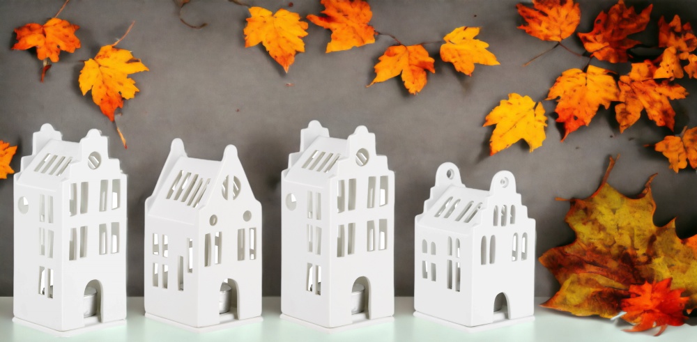 Vier weiße Lichthäuser im Grachtenhaus-Stil als Deko-Set mit herbstlichen Blättern im Hintergrund.