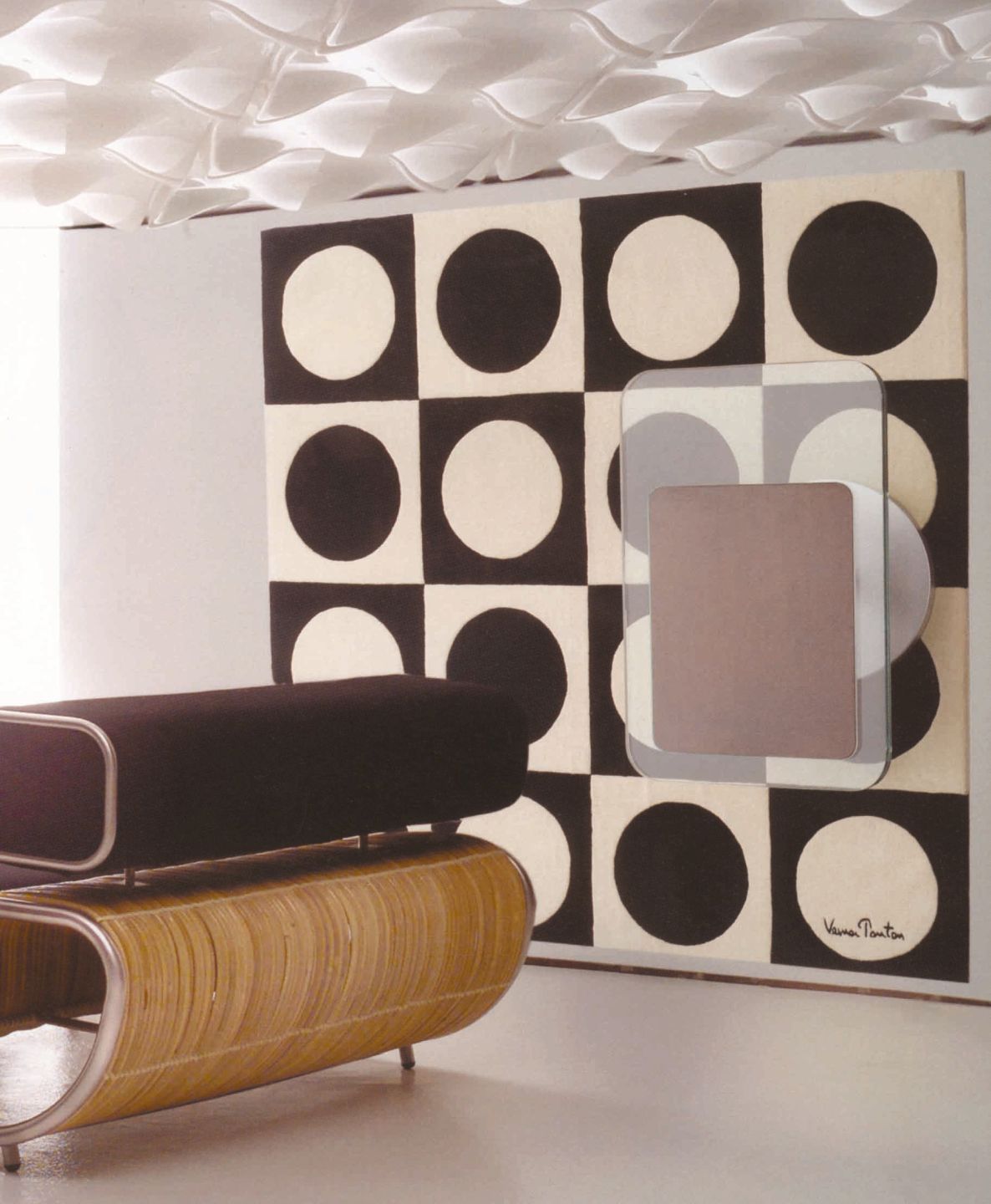 Braune Rattanliege mit schwarzer Polsterung vor schwarz-weißem Verner Panton Wandteppich und modernem Deckendesign.