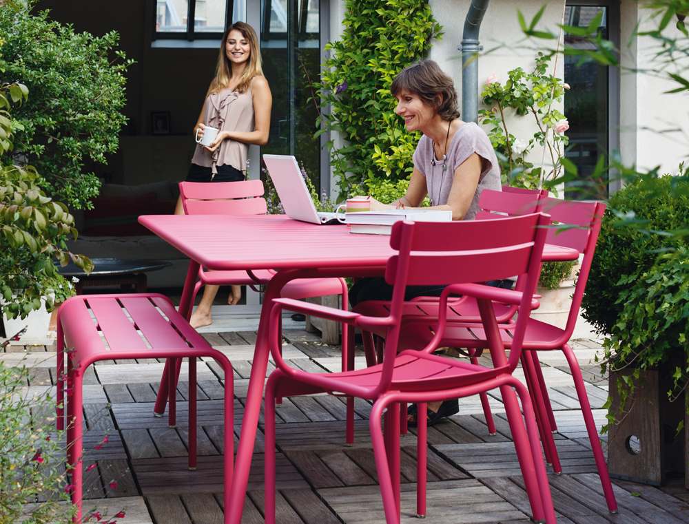Luxembourg Gartenmöbel-Set von Fermob in Pink: Tisch mit Stühlen und Hockern auf Holzterrasse mit zwei Frauen.