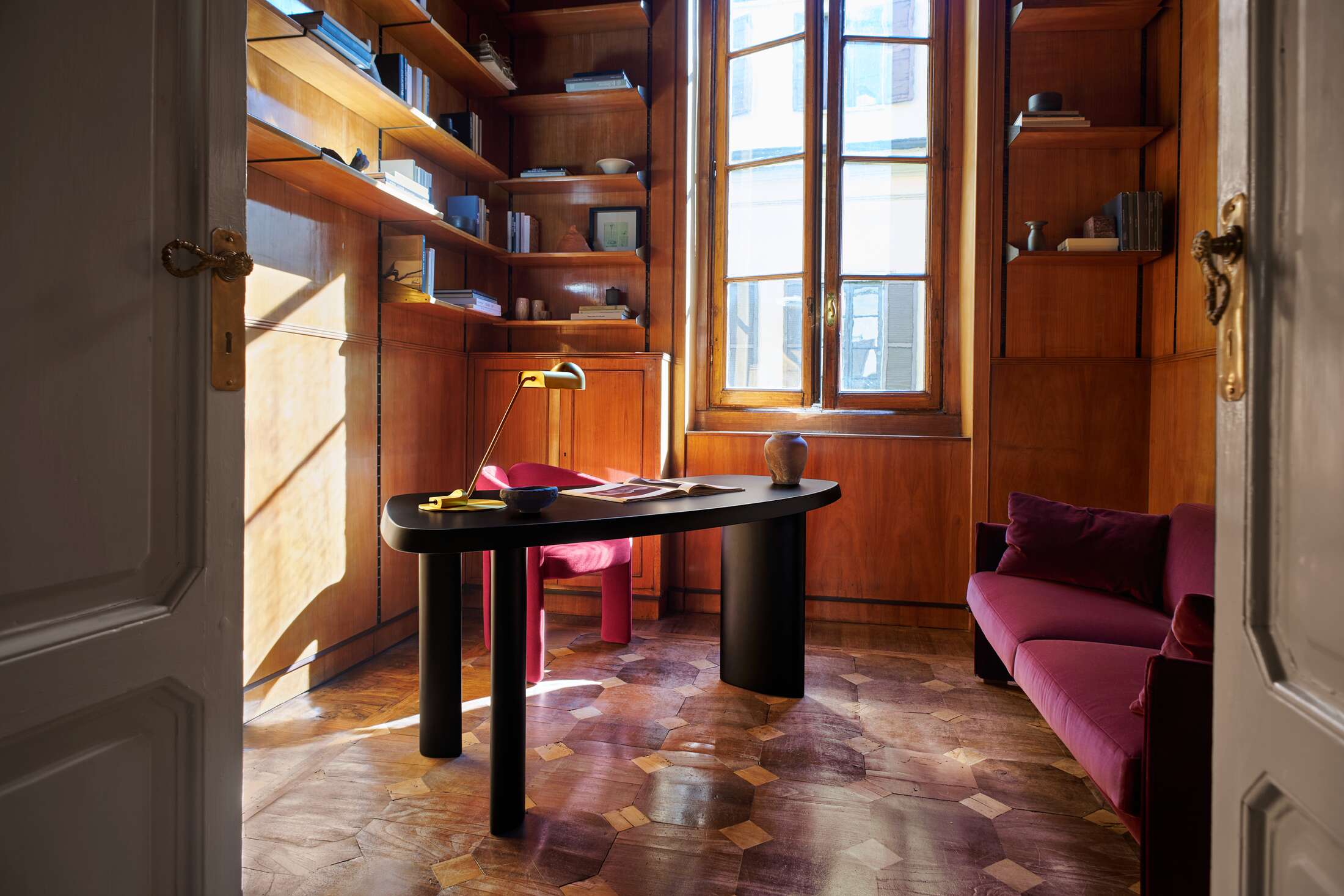 Innenaufnahme mit Cassina Dudet Sessel in Pink, schwarzem Schreibtisch und Sofa in einem holzgetäfelten Arbeitszimmer.