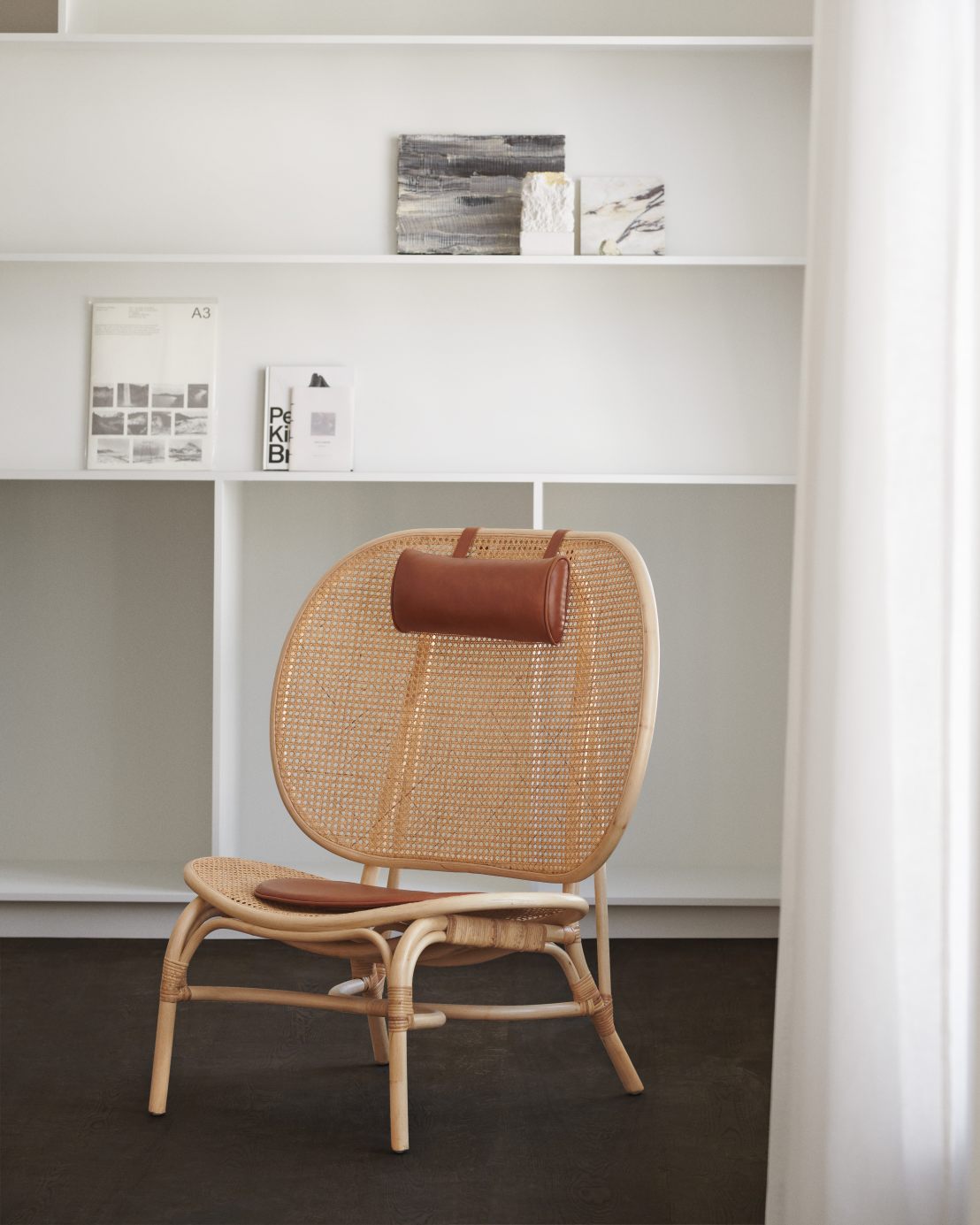 NORR11 Nomad Chair Sessel aus Rattan mit braunem Lederkissen vor weißem Regal.