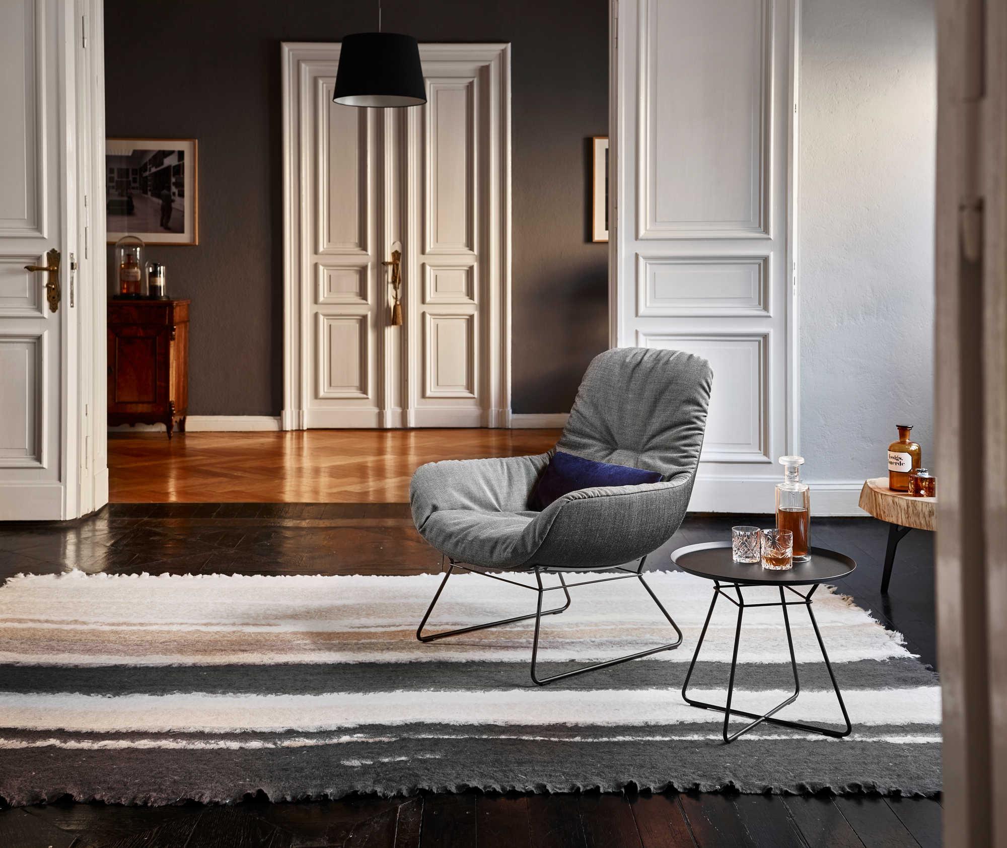 Grauer Leya Cocktail Loungesessel Freifrau Manufaktur mit Kissen und Beistelltisch in stilvollem Interieur.