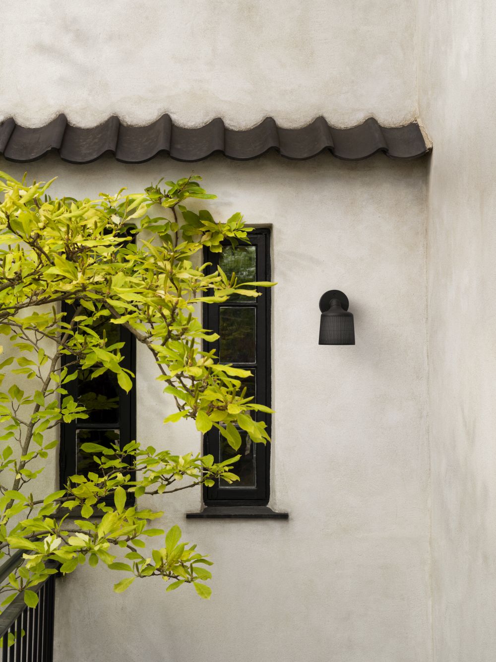 Schwarzer Vipp Outdoor Wandspot an einer weißen Hauswand neben einem Fenster und einem Baum.