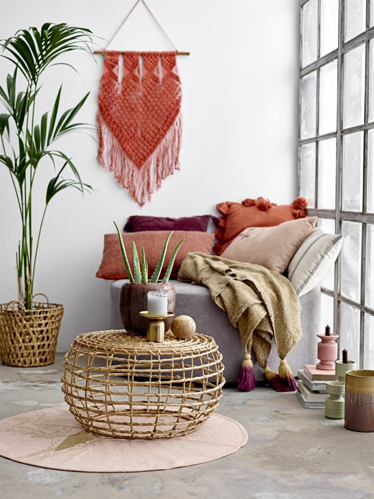 Wohnzimmer-Szene mit Rattan-Tisch, Kissen, Decke, Makramee-Wandbehang und Zimmerpflanzen für gemütliche Boho-Dekoration.