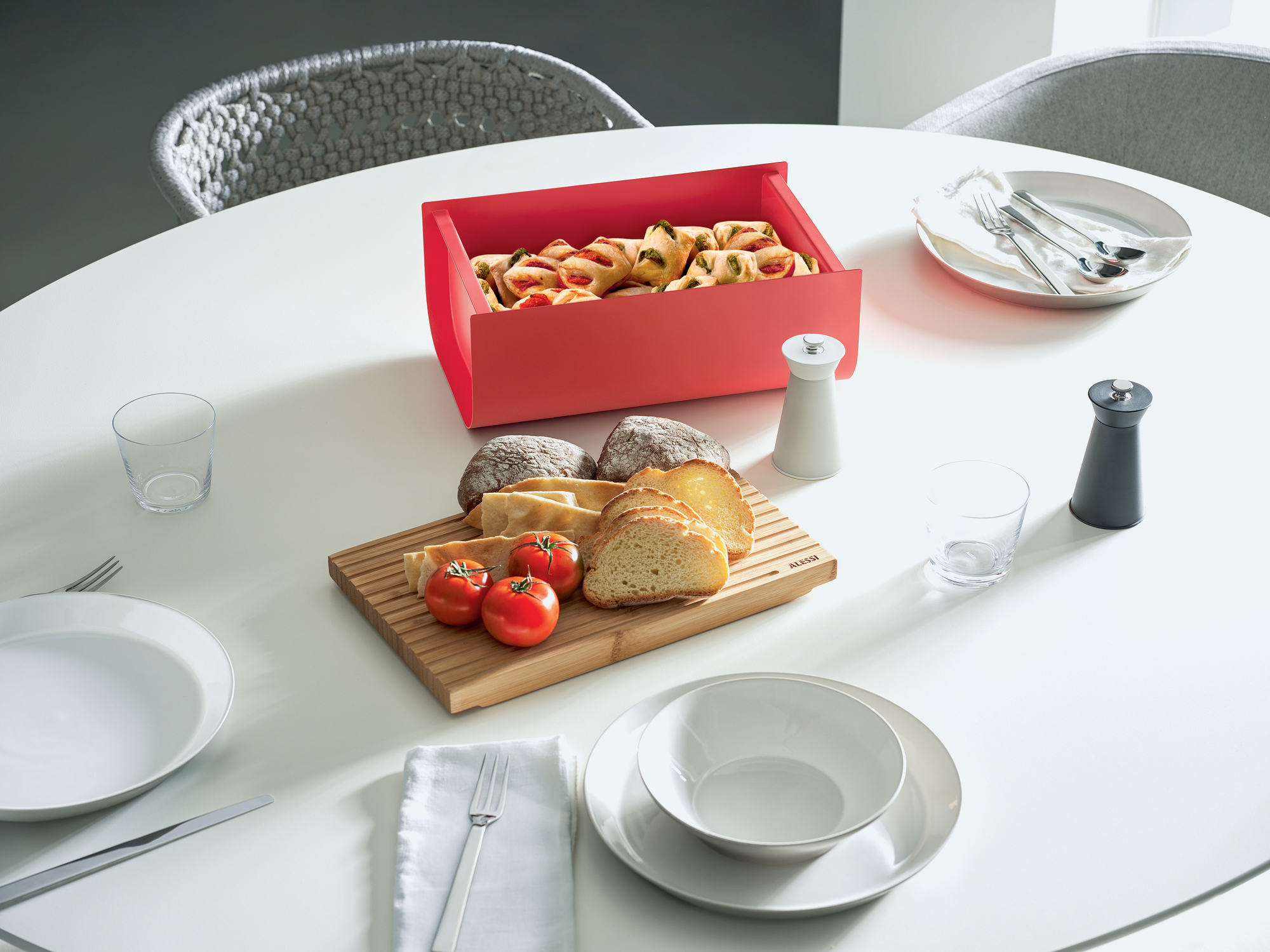 Roter Mattina Brotkasten von Alessi auf einem gedeckten Tisch mit Brot, Tomaten und Gebäck.