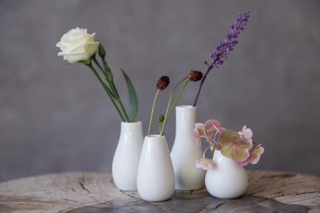 Produktfoto: Vier weiße Keramikvasen mit verschiedenen Blumen auf einem Holztisch.