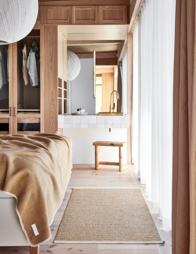 Heller Schlafzimmerbereich mit beigem Teppich, Holzhocker, Bett mit Decke und Einbauschrank im natürlichen Design.